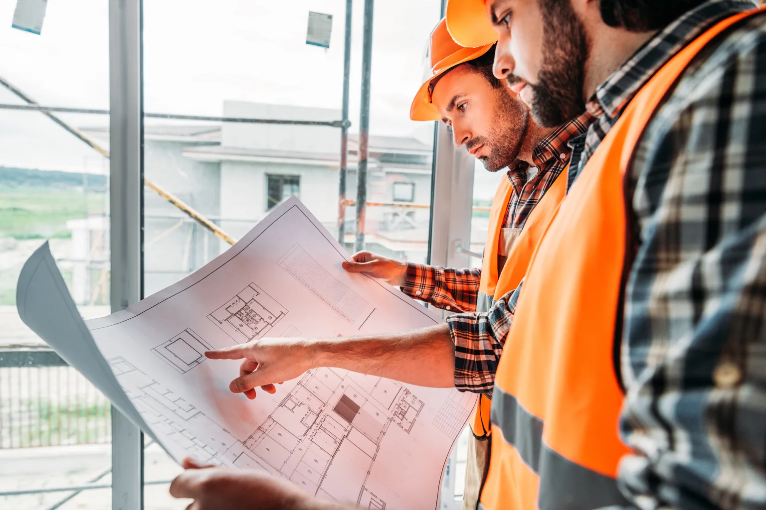 Two builders in hard hats and safety vests reviewing a printed blueprint together at a window on a job site, one pointing to a section of the plan.