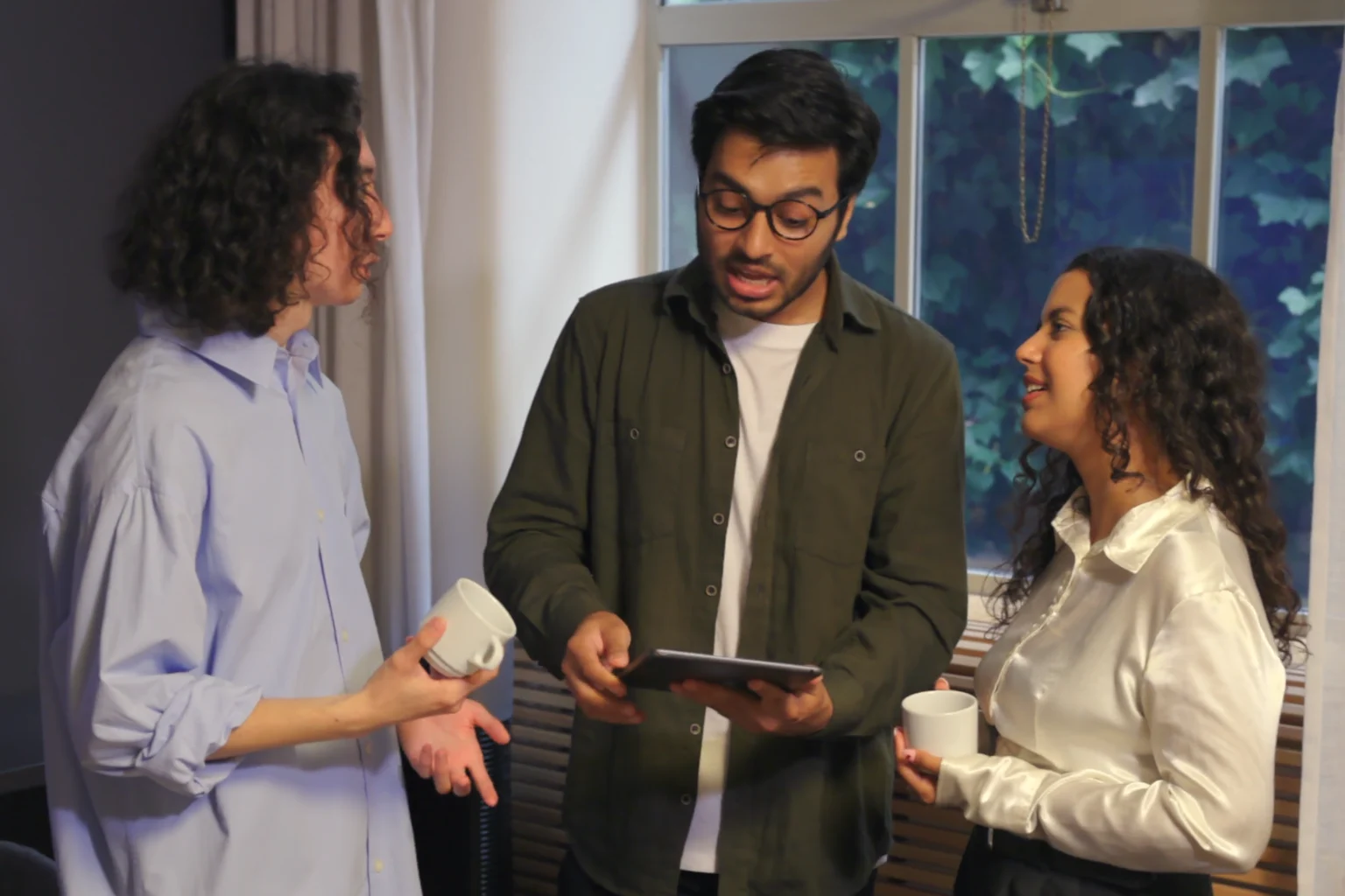 Three colleagues standing by a window with coffee, one walking the others through work on a tablet.