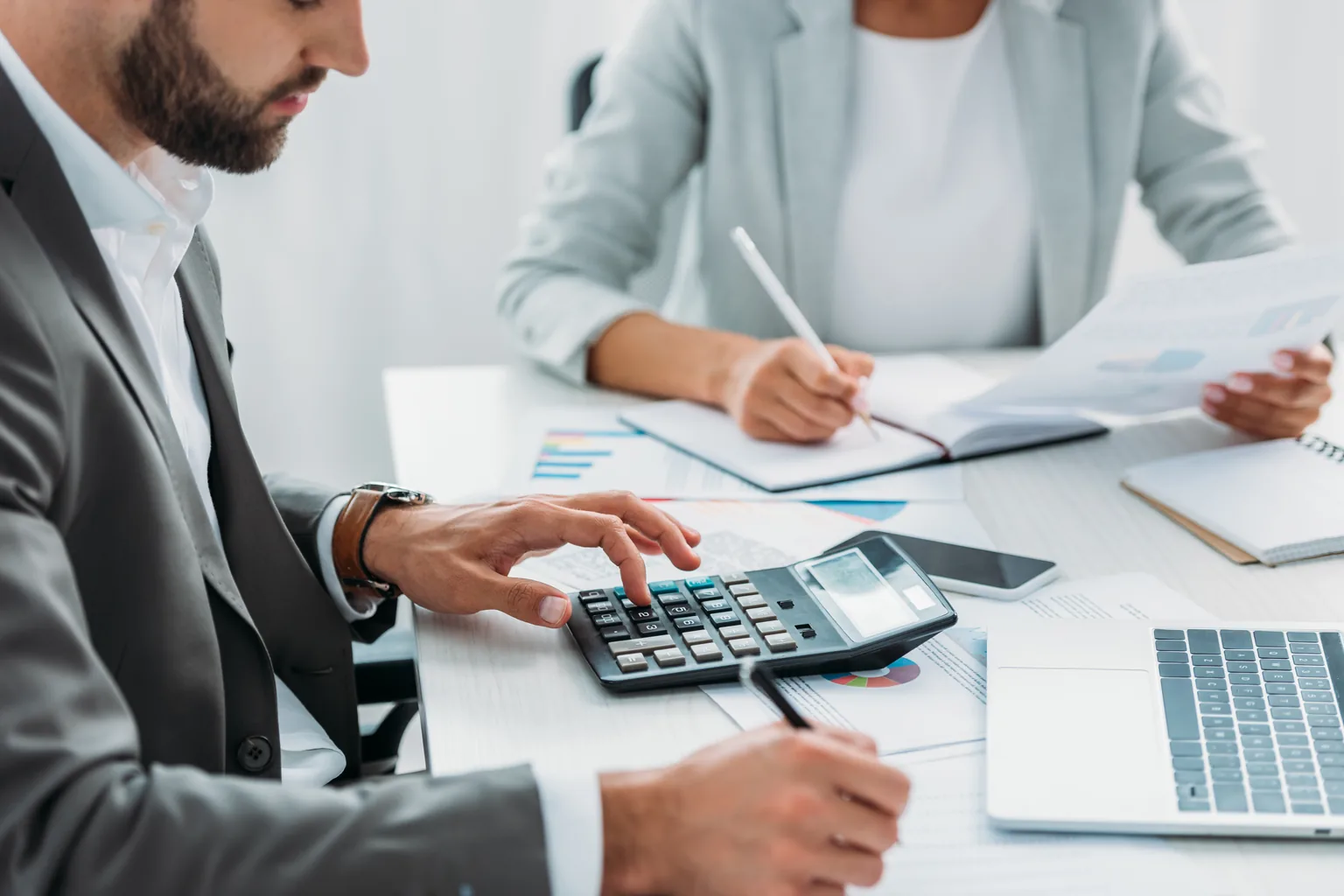 Two colleagues at a white desk working through figures, one entering numbers on a calculator while the other takes notes from a printed report.